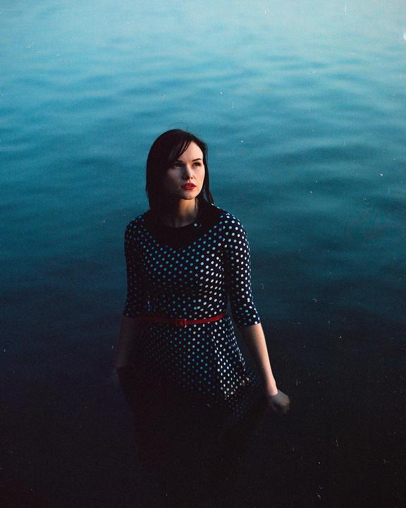 A woman in a polka dot dress with a red belt stands in dark water.
