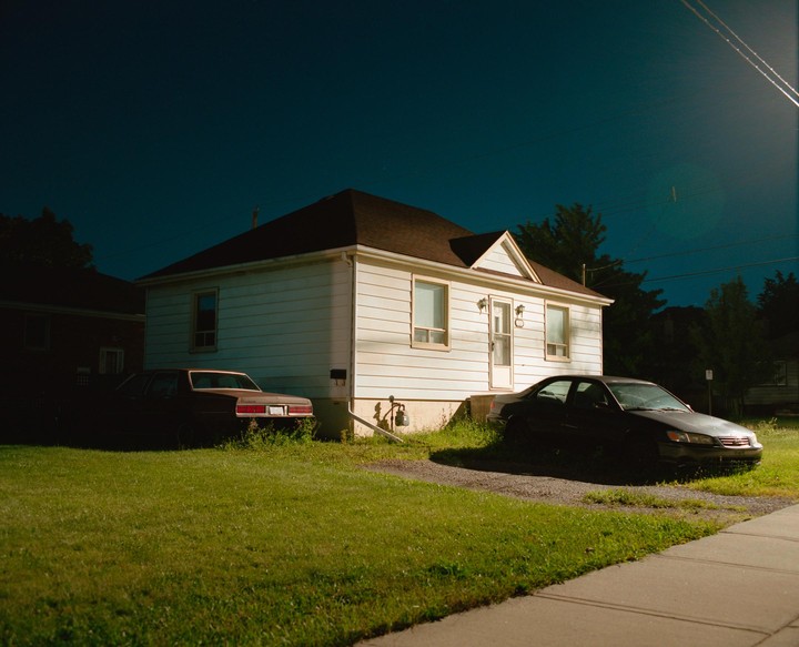 Two cars are parked in front of a white house with a brown roof at night.