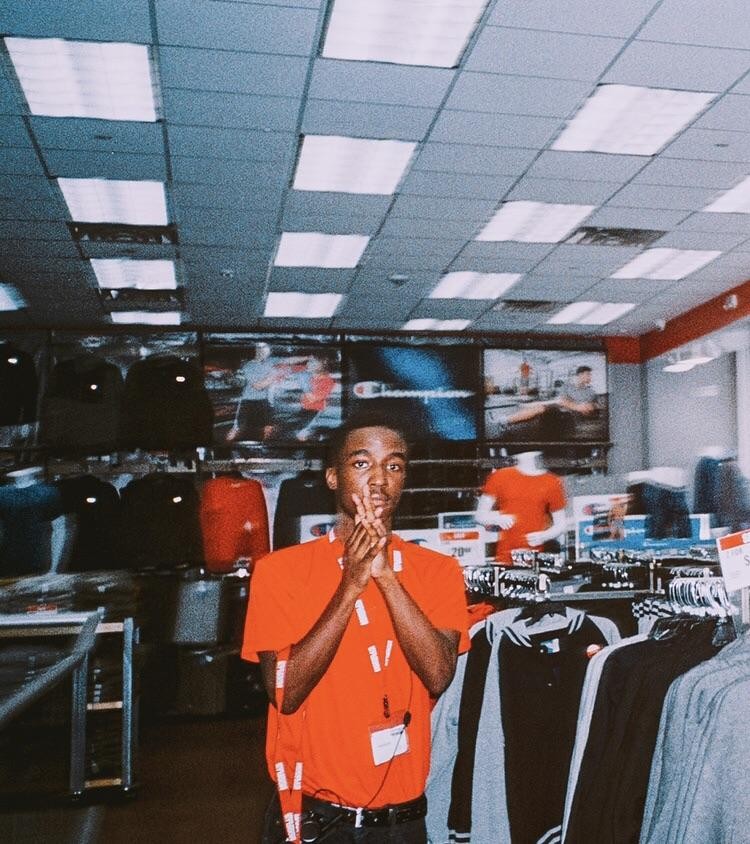 A man in an orange shirt holds his hands up in a peace sign gesture while standing in a clothing store.