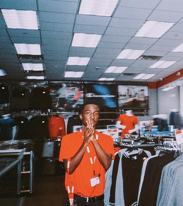 A man in an orange shirt holds his hands up in a peace sign gesture while standing in a clothing store.