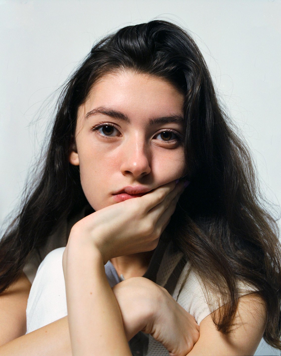 A woman with long dark hair rests her chin on her hand while looking directly at the viewer.