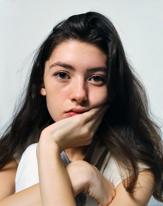 A woman with long dark hair rests her chin on her hand while looking directly at the viewer.