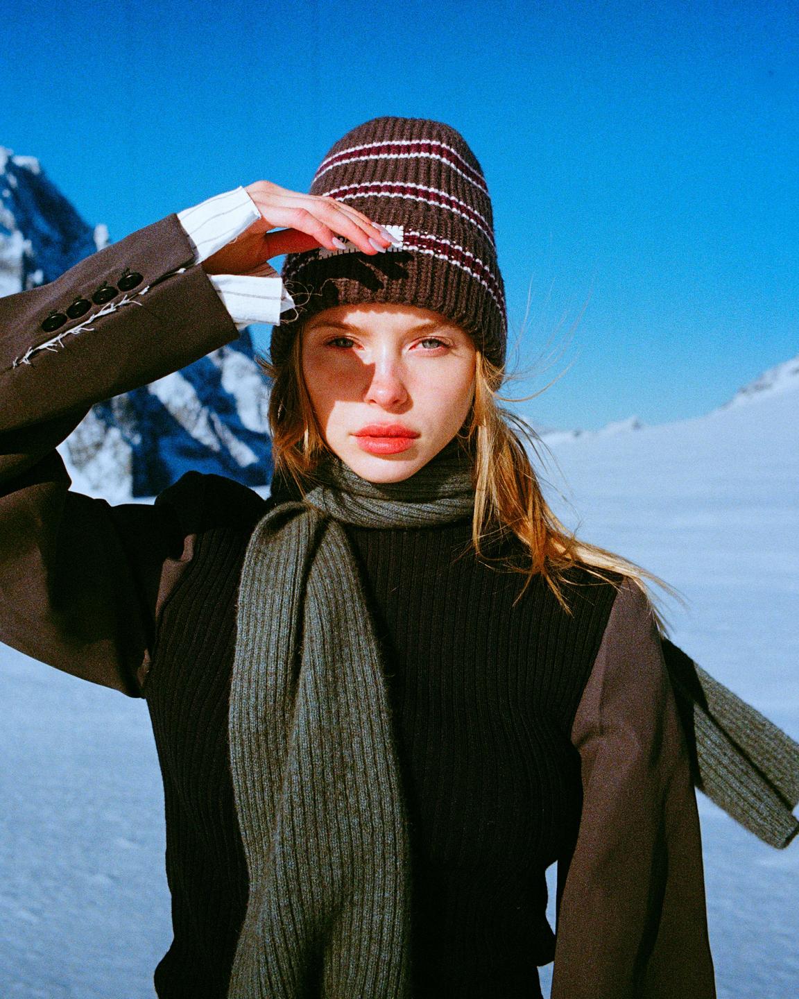 A woman wearing a beanie and scarf shields her eyes from the sun in a snowy mountain landscape.