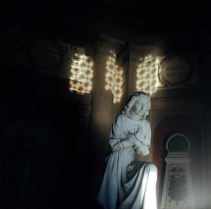 A statue of a kneeling woman with her hands clasped to her chest is illuminated by dappled light.