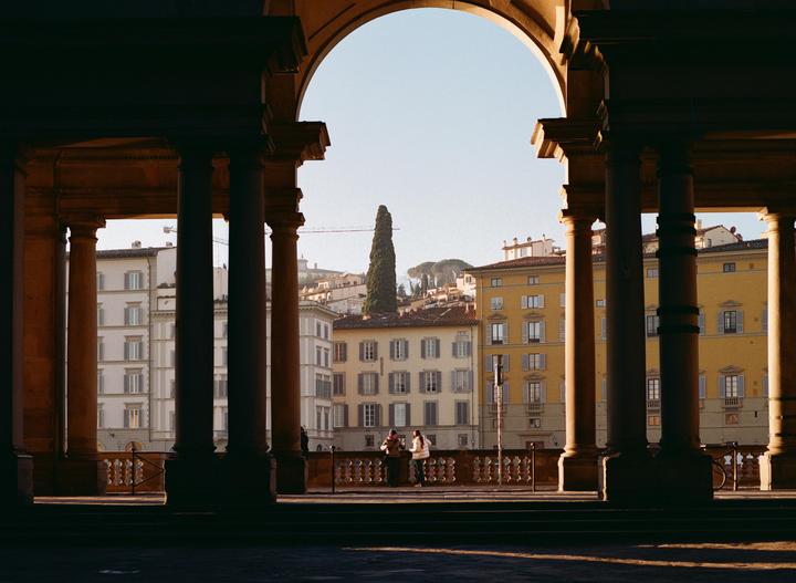 Two women stand on a balcony overlooking a city with buildings and a tall tree, framed by an archway and columns.