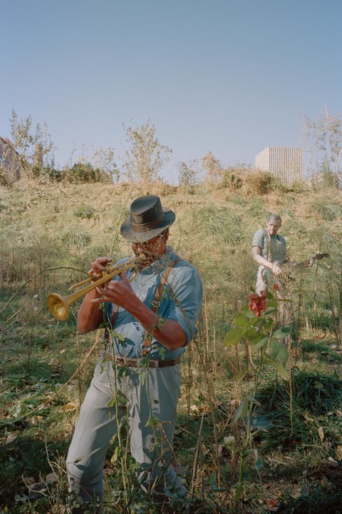 A man in a hat plays a trumpet in a field with another man in the background.