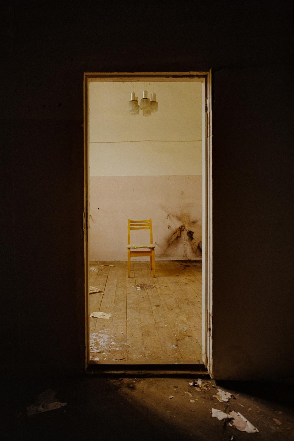 A yellow chair sits in a dilapidated room with a chandelier visible through an open doorway.