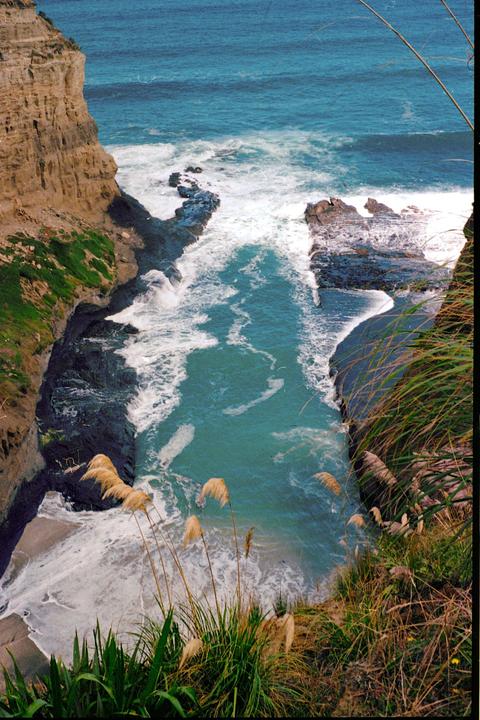 A narrow inlet of turquoise water with white waves crashes against rocky cliffs and a small sandy beach.