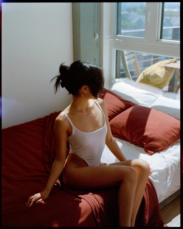 A woman sits on a bed next to a window, looking out.