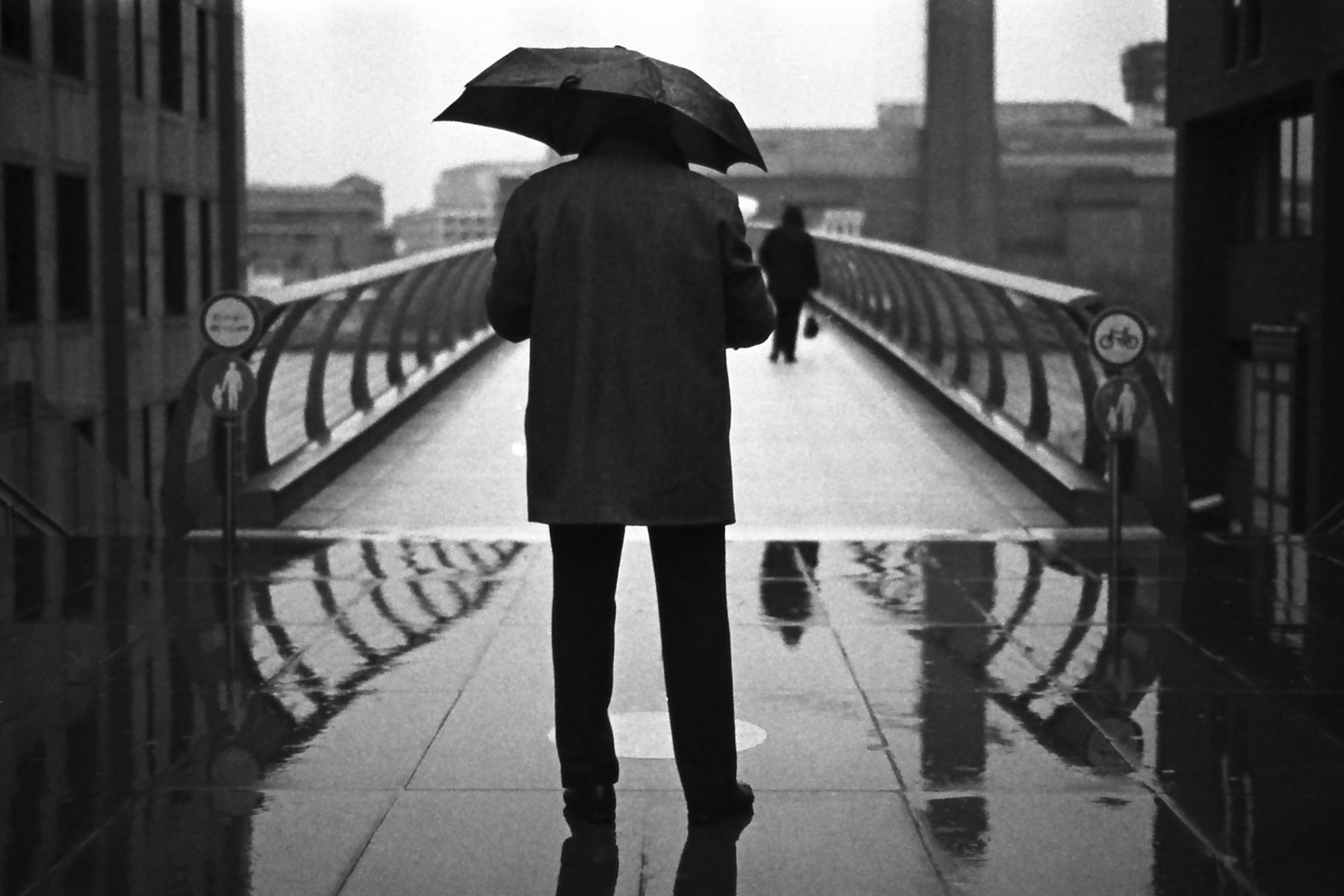 A person holding an umbrella stands on a wet bridge with another person in the distance.