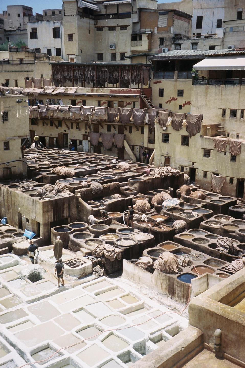 A high-angle view shows a tannery with many stone vats filled with colorful liquids, surrounded by buildings with hides drying on their roofs.