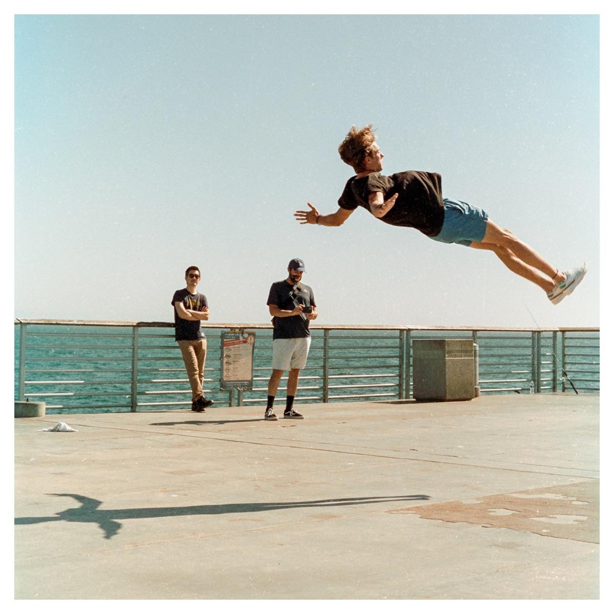 A man appears to float in the air above a pier while two other men stand nearby.