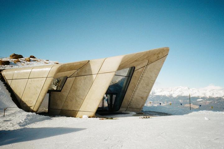 A modern building with angular concrete walls and large windows stands in a snowy landscape with mountains in the background.