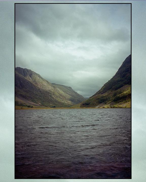 A body of water with ripples on its surface is surrounded by mountains under a cloudy sky.