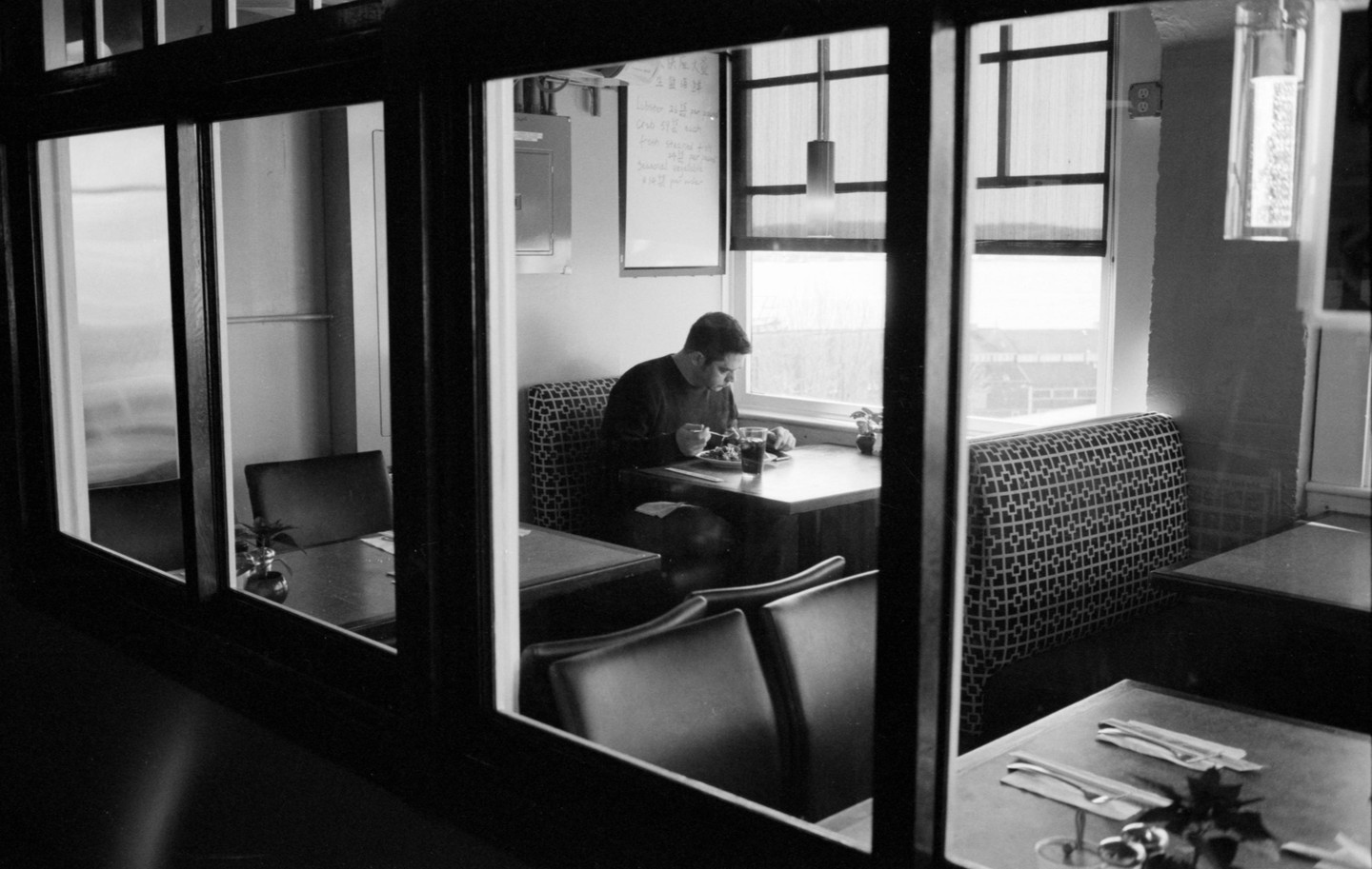 A man eats alone at a table in a diner, viewed through a window.