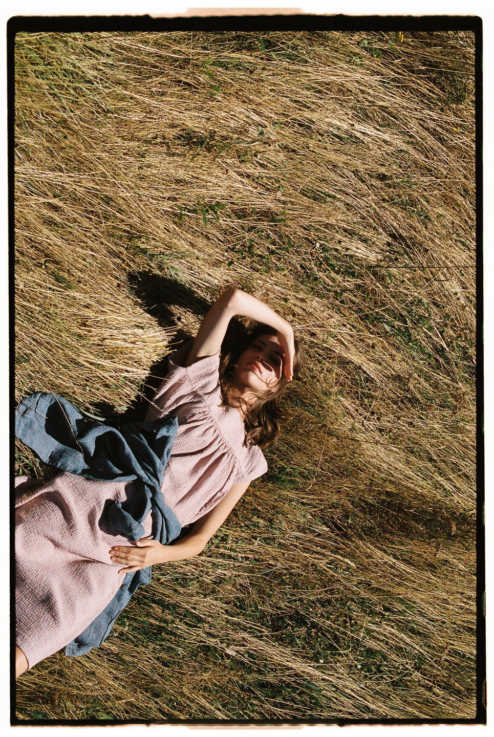 A woman in a pink dress and blue fabric lies in dry grass, shielding her eyes from the sun.