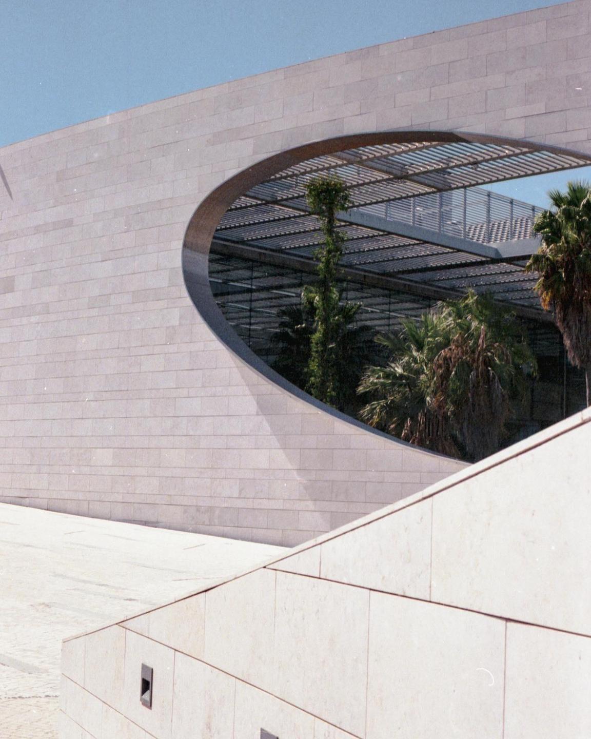 A modern building with a large oval opening reveals palm trees and a metal canopy inside.