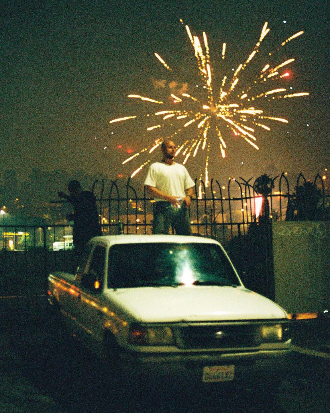 A man stands in front of a fence with a white pickup truck in the foreground and fireworks exploding in the night sky.