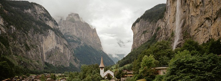 A small village with a church is nestled in a valley between two towering mountains with waterfalls.