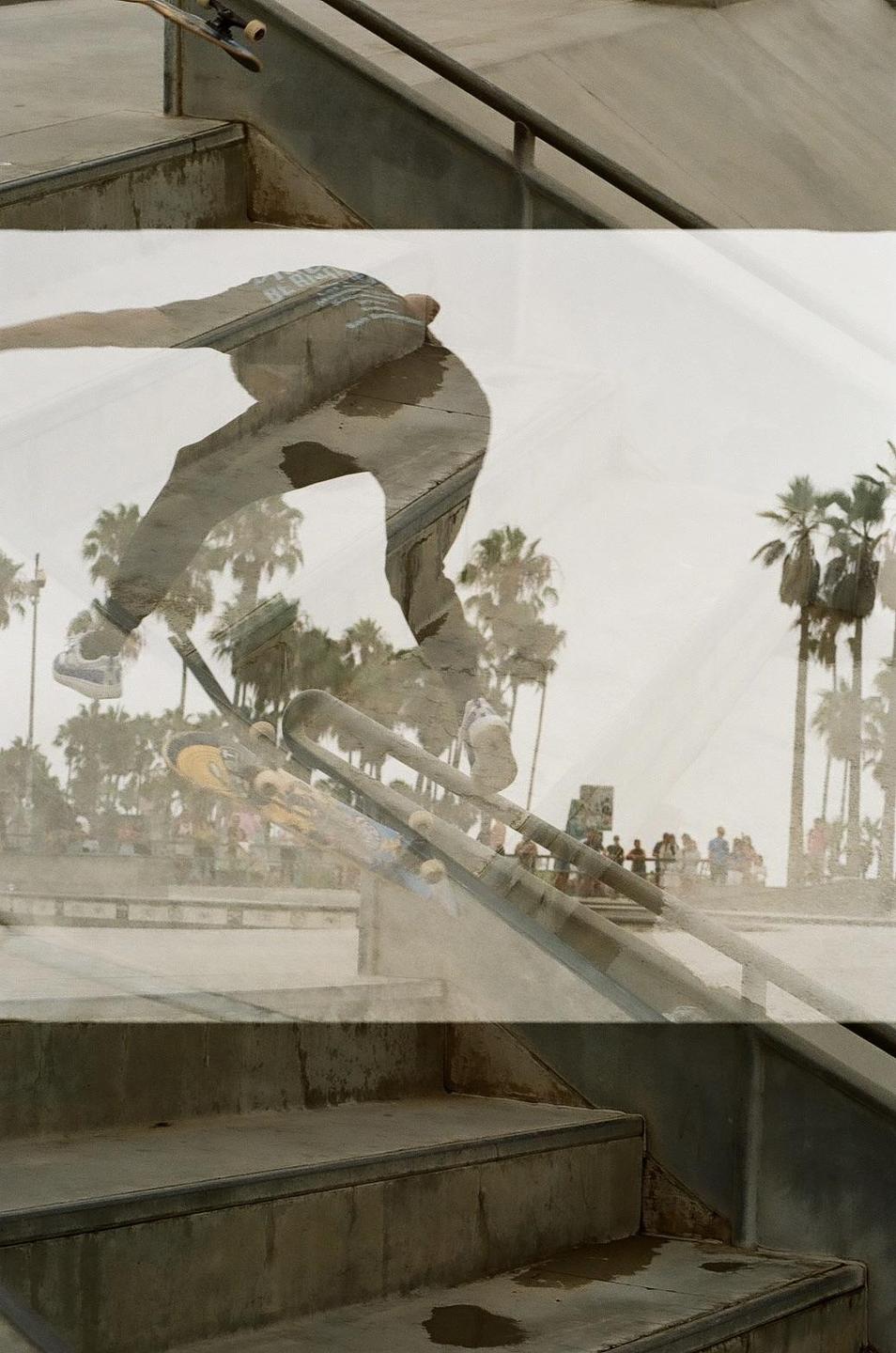 A skateboarder grinds a rail at a skate park with palm trees and people in the background.