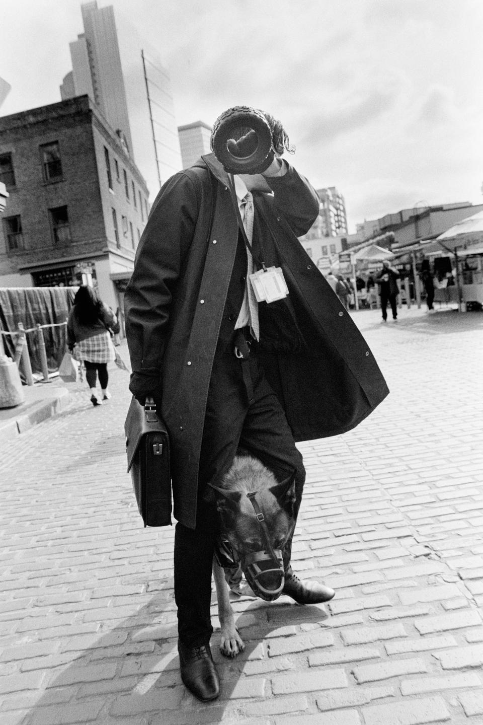 A man in a trench coat holds a briefcase and a dog with a muzzle stands between his legs on a brick street.