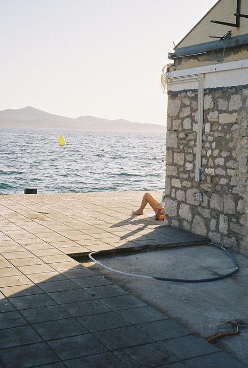 A person is relaxing on a tiled pier next to a stone building, with the sea and mountains in the background.