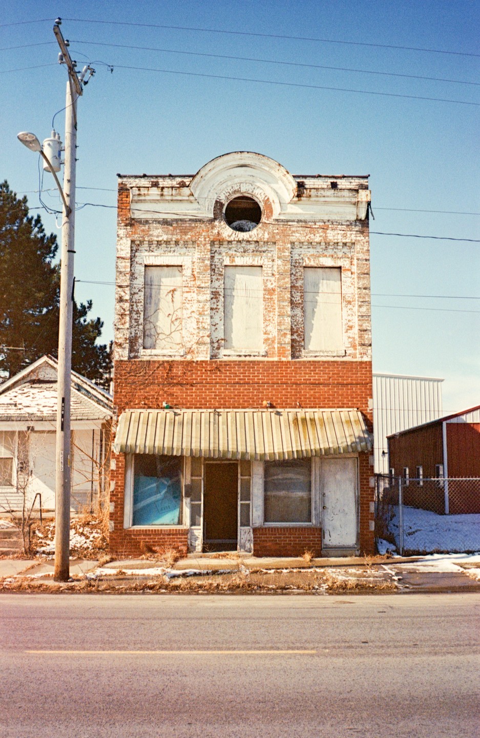 An old brick building with boarded-up windows and a striped awning stands beside a street and a utility pole.