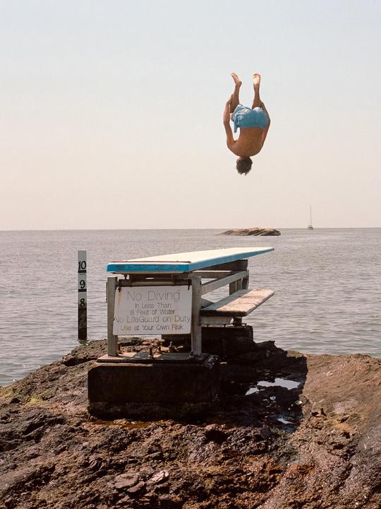 A man performs a backflip off a diving board over the ocean, with a "No Diving" sign visible on the board's support structure.