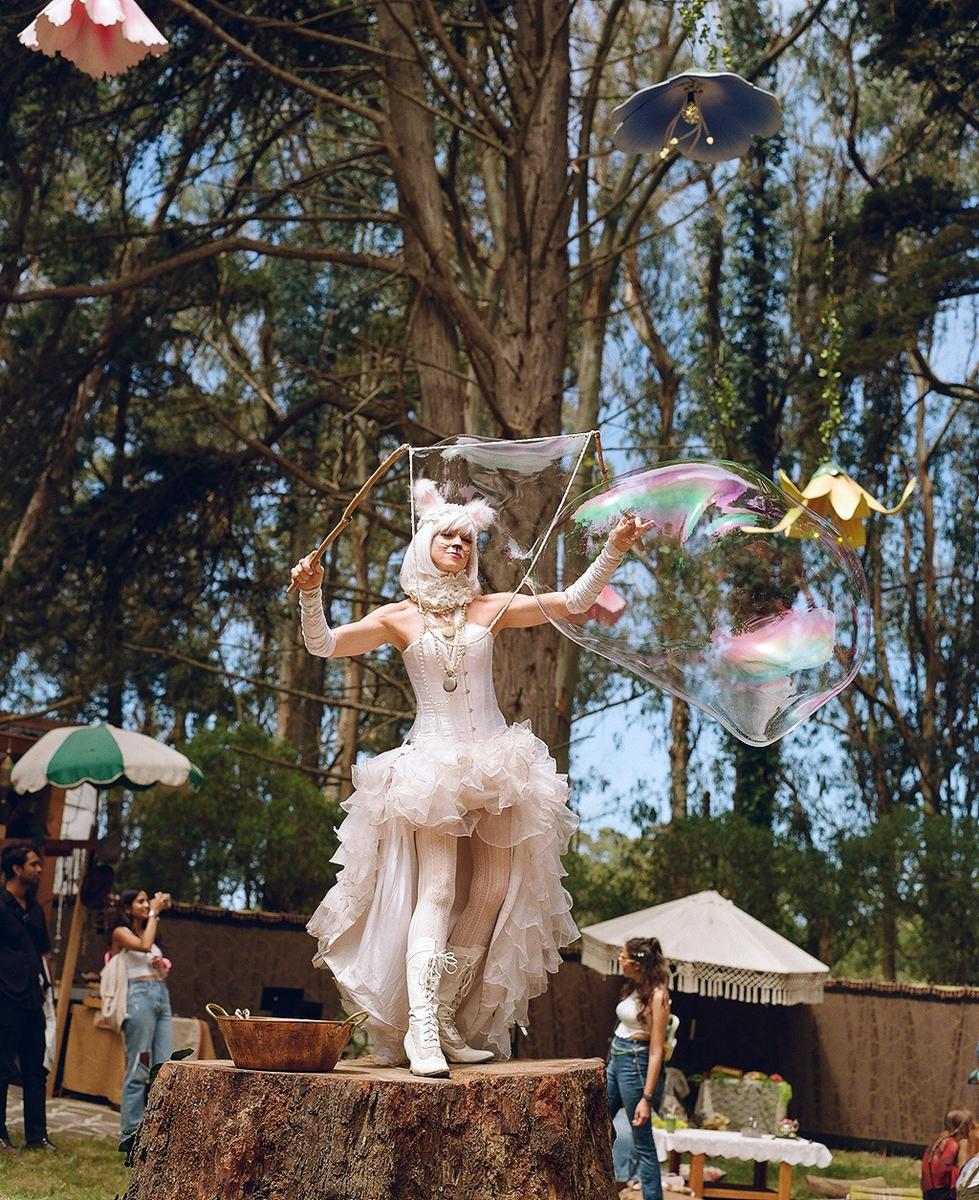 A woman in a white costume with bunny ears stands on a tree stump and blows a large bubble.