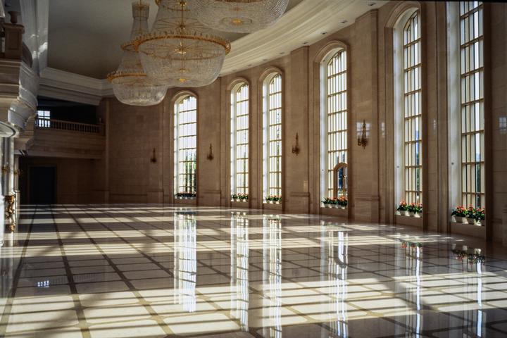Sunlight streams through tall arched windows, casting long shadows and reflections on a polished marble floor in a grand hall.