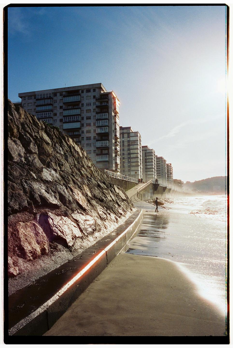 A person carrying a surfboard walks along a wet beach next to a rock wall and several tall buildings under a bright sky.