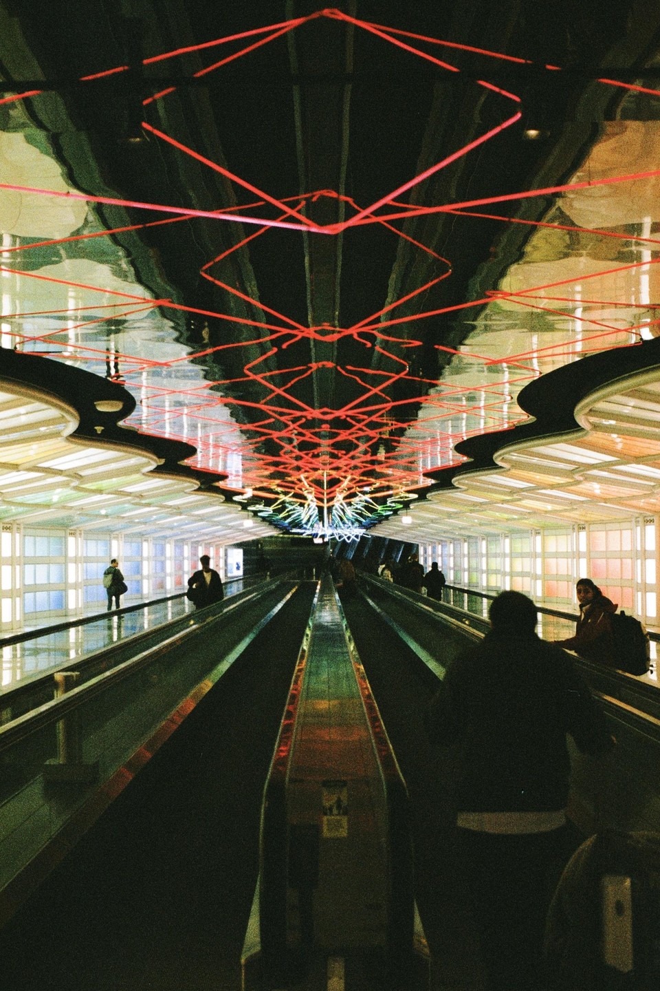 People ride moving walkways under a ceiling with red neon lights and colorful panels.