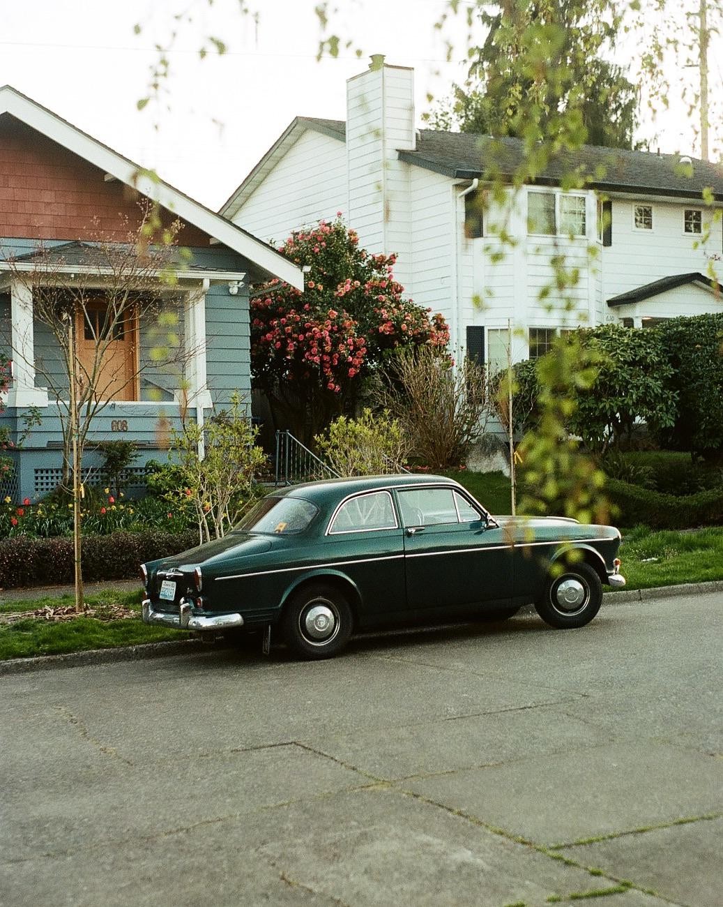 A dark green vintage car is parked on a residential street in front of houses with blooming bushes.