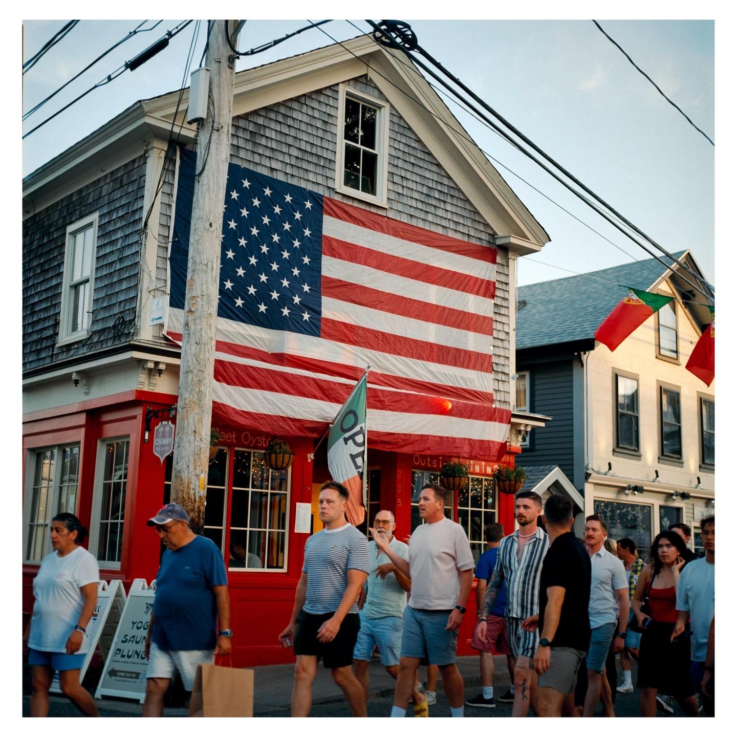 A large American flag hangs on the side of a red building with people walking on the street in front of it.