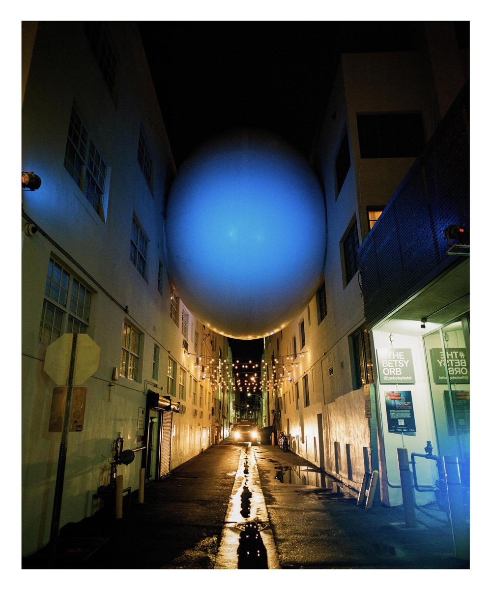 A large blue orb is suspended between two buildings above a wet street at night.