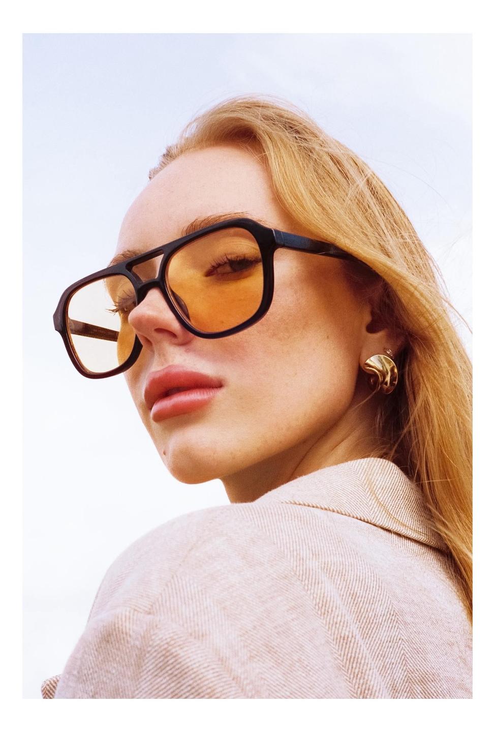 A woman with long hair and orange-tinted glasses looks over her shoulder.