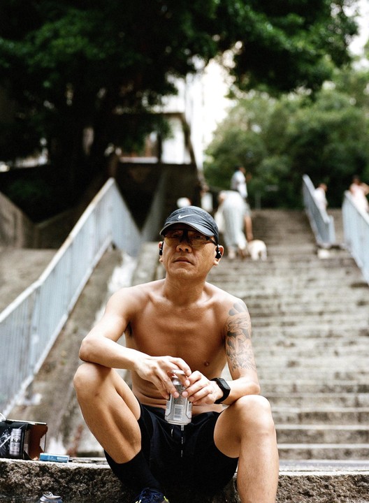 A man sits shirtless on concrete steps, holding a can and looking forward.