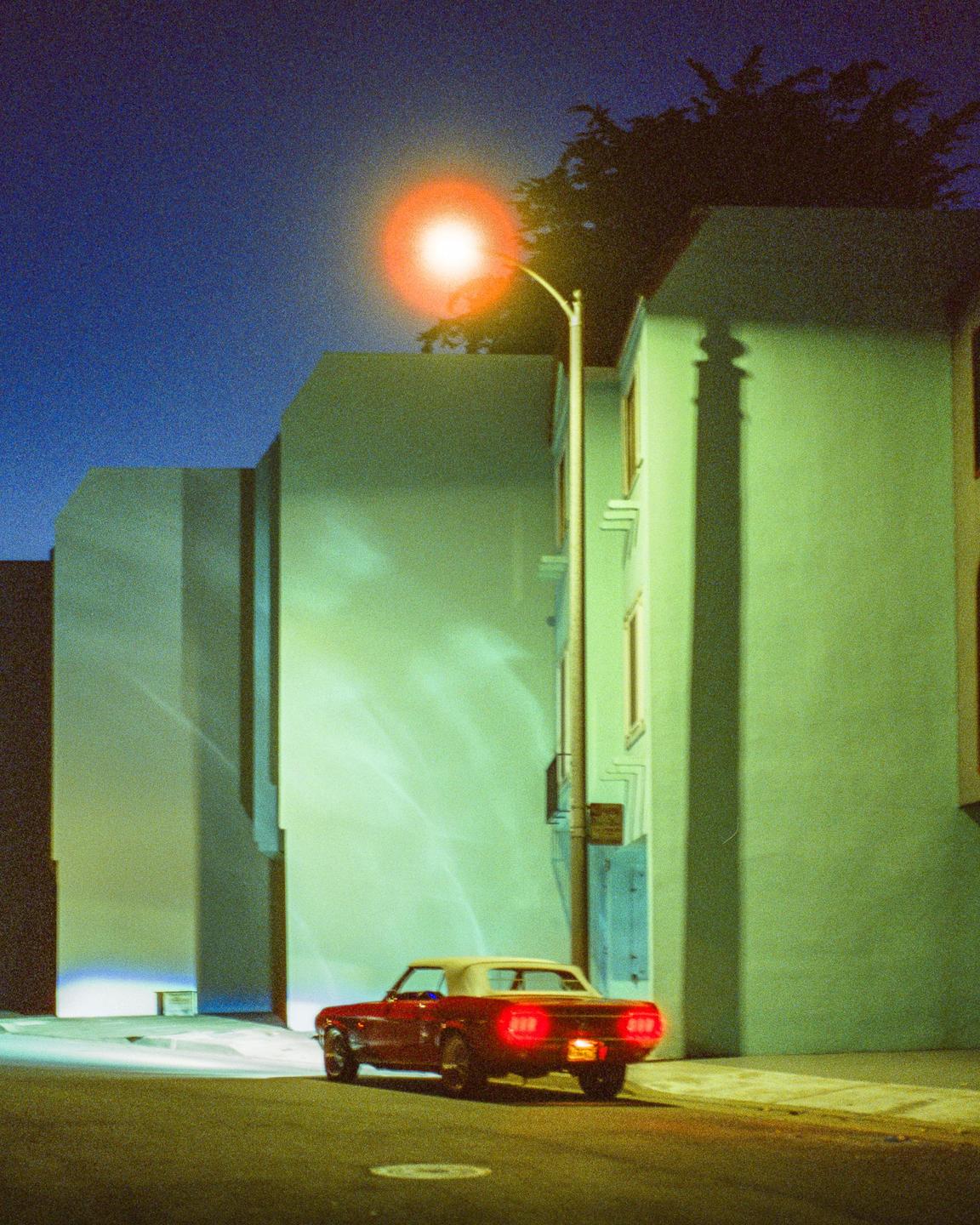 A red convertible car is parked on a street at night next to buildings with a bright street light overhead.