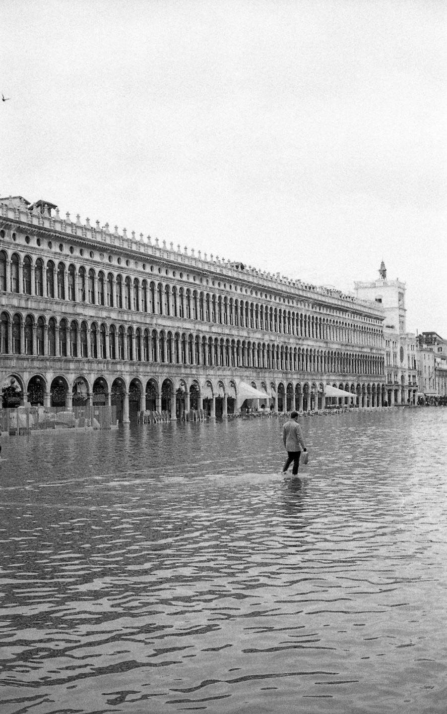 A man wades through floodwaters in front of a large building with many arched windows.