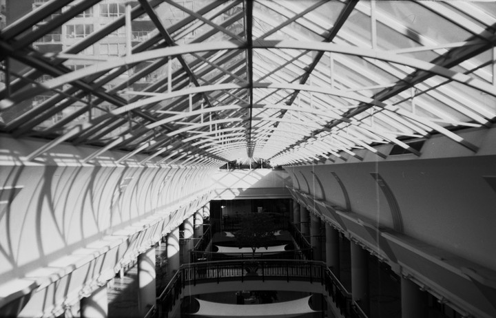A black and white photograph shows the interior of a building with a glass ceiling and strong shadows cast on the walls.