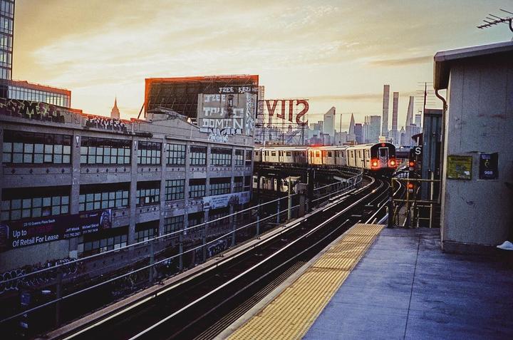 A subway train approaches a station platform with a city skyline in the background at dusk.