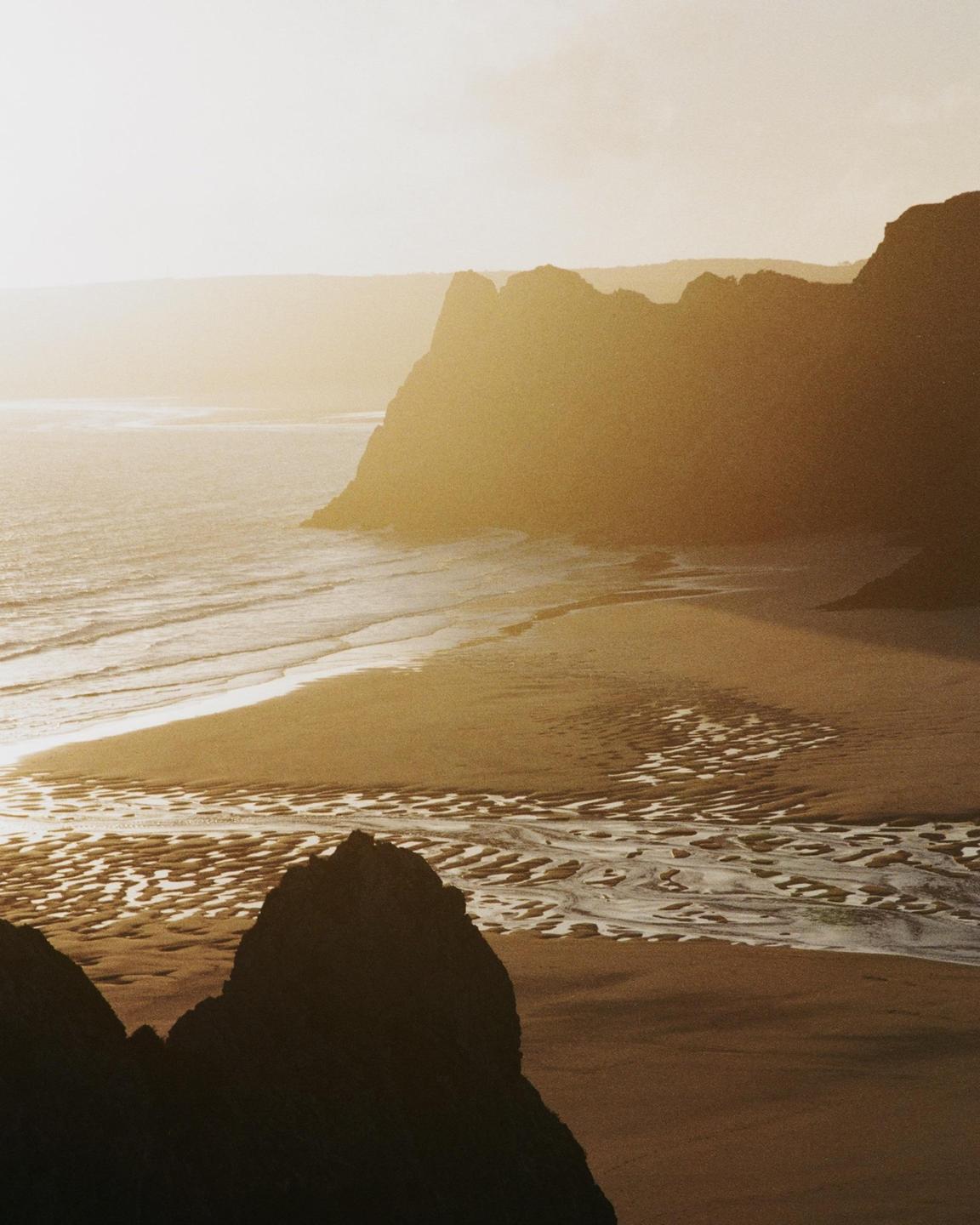 The sun shines over a beach with cliffs and ocean waves.