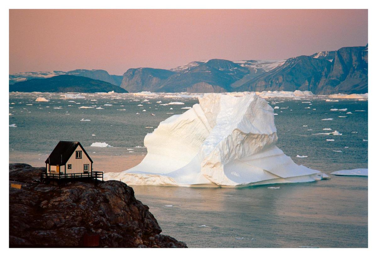 A small house sits on a rocky cliff overlooking a body of water filled with icebergs and surrounded by mountains under a pink sky.