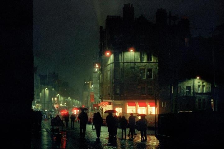 A street at night is filled with people holding umbrellas, with illuminated buildings and reflections on the wet ground.