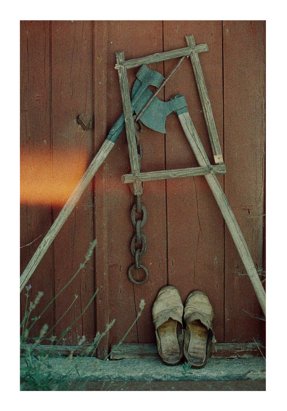 Two axes, a wooden frame, a metal chain, and a pair of worn slippers lean against a red wooden wall.