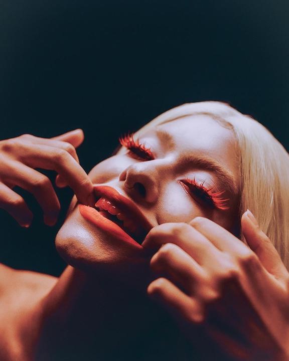 A woman with red eyelashes and red lipstick pulls at her lips with her fingers.