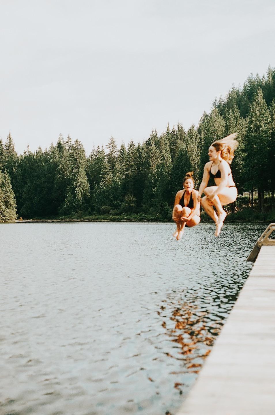 Two women jump into a lake from a wooden dock surrounded by a forest.