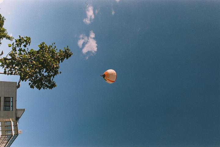 A hot air balloon floats in a clear blue sky above a building and tree.