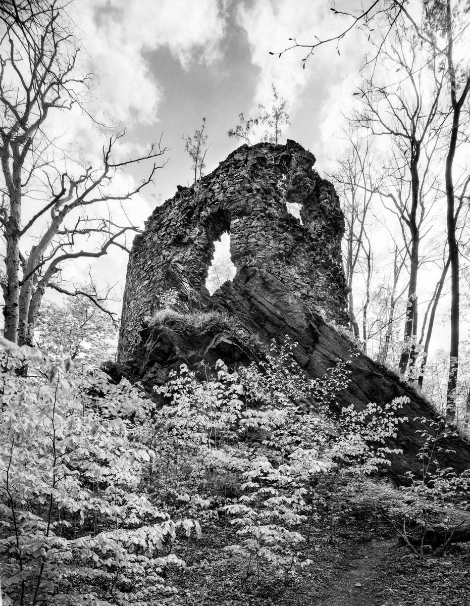 A stone ruin stands on a hill surrounded by trees and foliage under a cloudy sky.