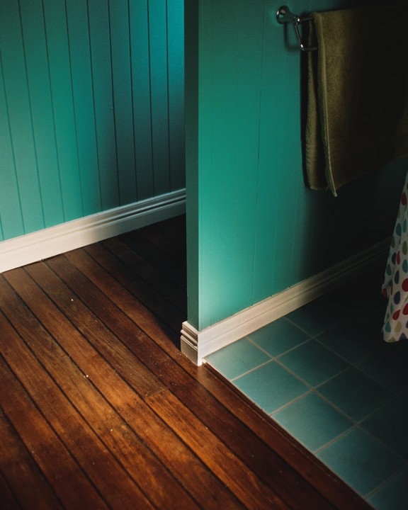 A teal wall with white trim meets a wooden floor on the left and teal tiled floor on the right, with a towel hanging on the wall.